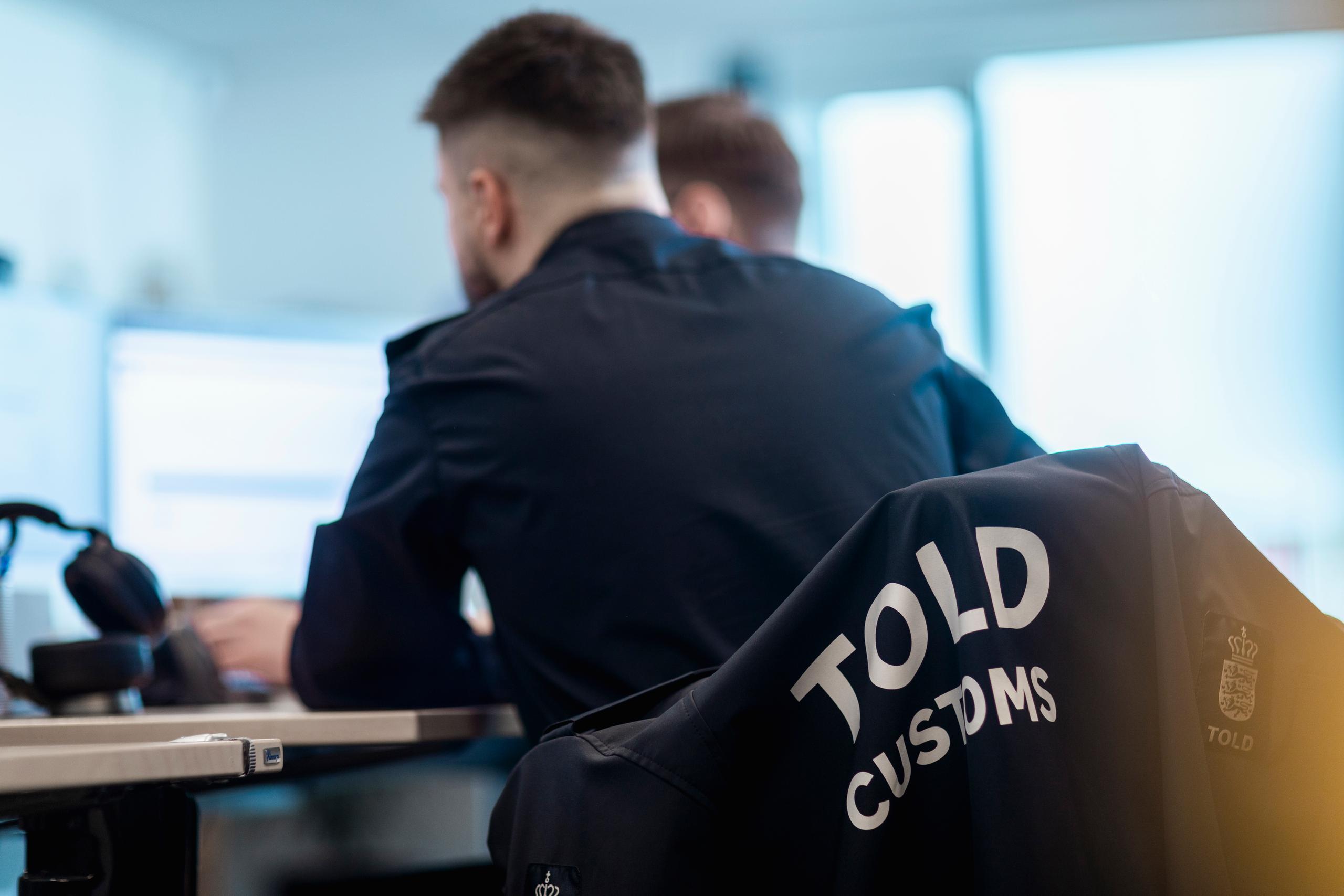 Tolder som sidder foran en computer. Man kan se ryggen af ham, og på stolen hænger der en toldjakke