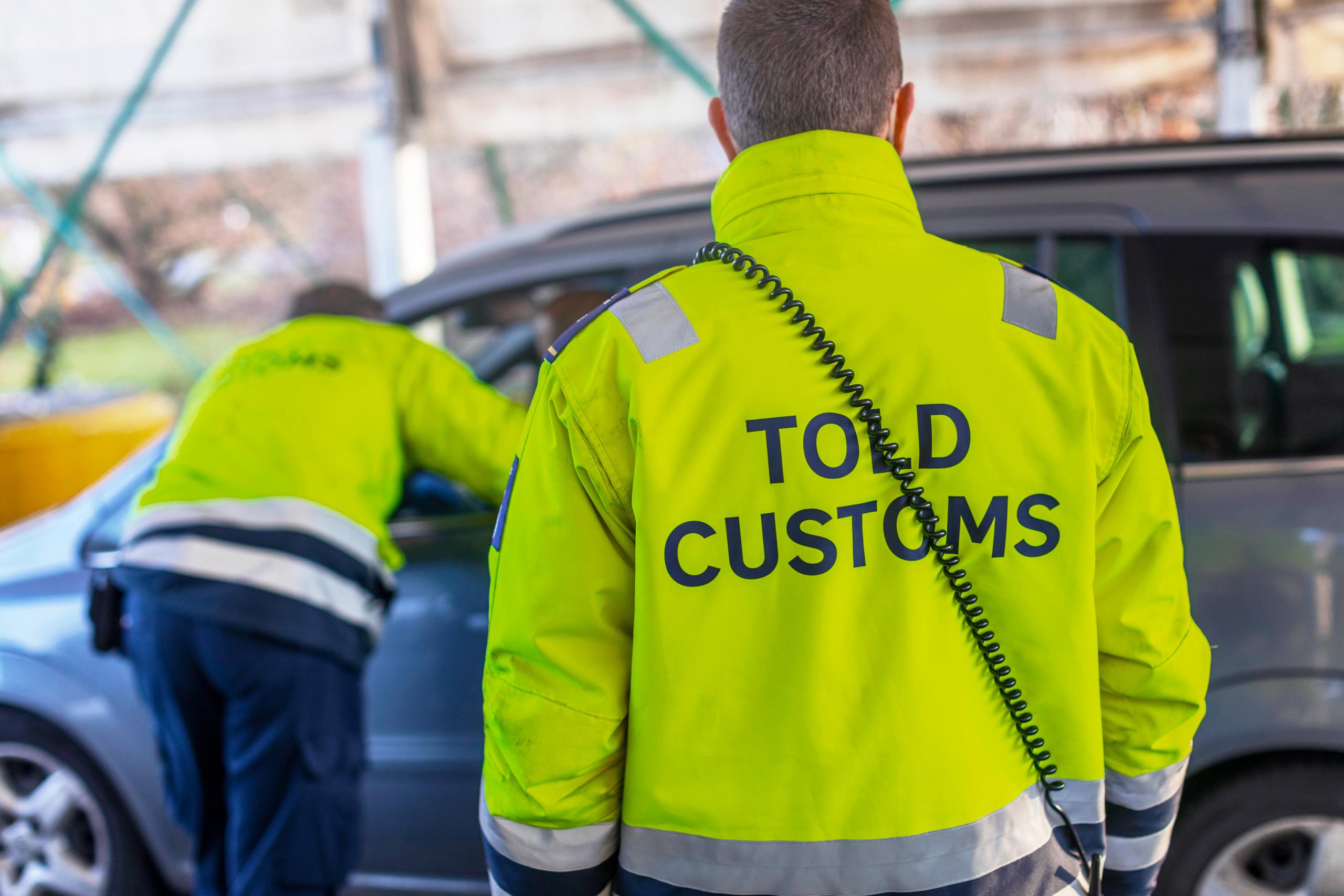 Toldkontrol i Padborg. Ryggen af to toldere, med toldjakker, den ene taler med person i bil
